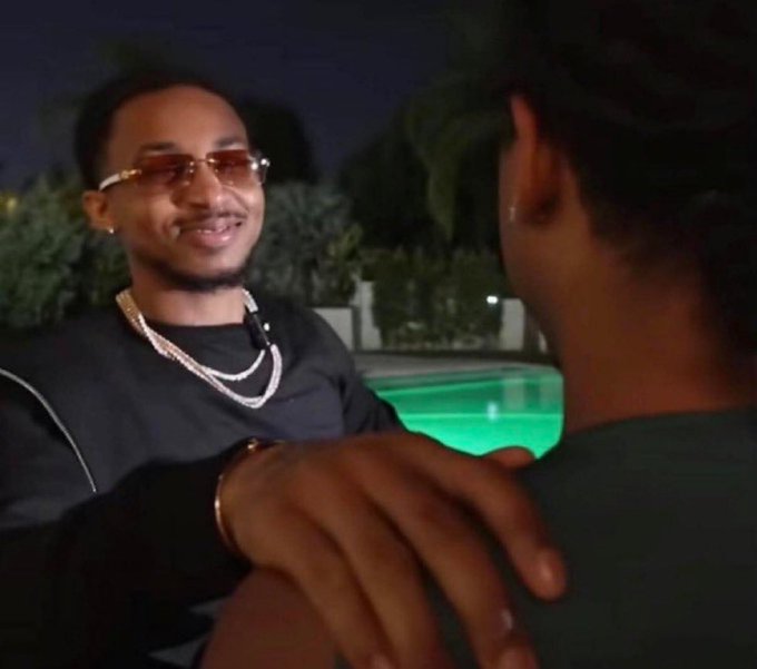 A smiling man wearing sunglasses, diamond chains, and a black outfit places his hand on another person's shoulder at night by a pool, appearing to greet them warmly. The image captures a friendly, celebratory moment that contrasts with the post's skeptical tone about someone pivoting to NFT trading as a newcomer to the space.