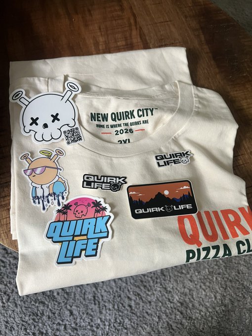 A folded white t-shirt with "NEW QUIRK CITY" text surrounded by various "Quirk Life" themed stickers, placed on a wooden table.