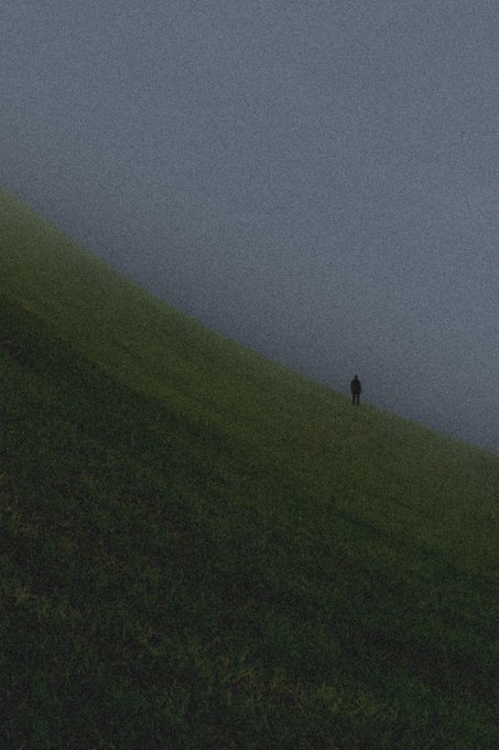 A solitary figure stands on a green grassy slope against a vast, moody gray-blue background.