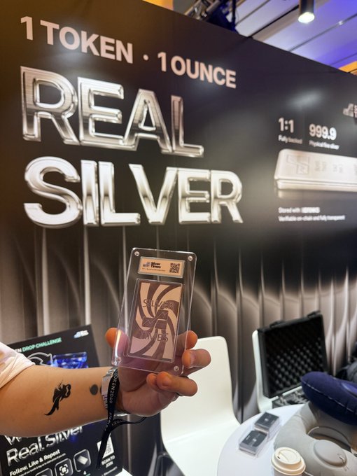 A hand holds a silver bar encased in a protective sleeve in front of a large advertisement for 'REAL SILVER' with the text '1 TOKEN · 1 OUNCE'.