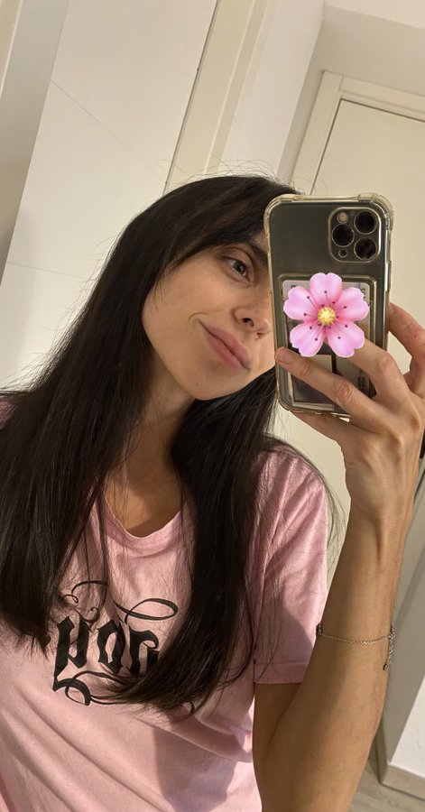 A woman with long dark hair takes a mirror selfie wearing a pink t-shirt, holding an iPhone with a pink cherry blossom decoration on the case.