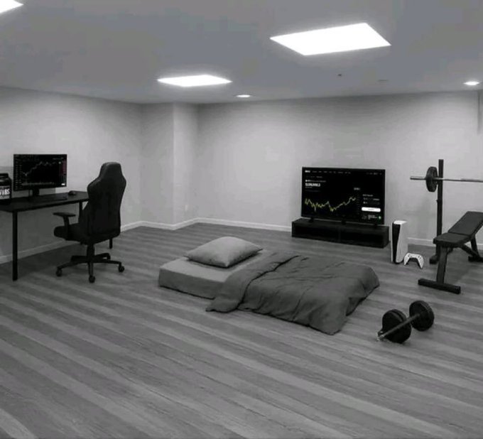 A minimalist bedroom/gaming room with a bed on the floor, a gaming chair at a desk with a monitor showing stock charts, and a weight bench and dumbbell nearby.