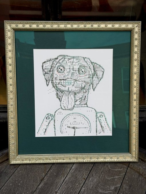 A framed black-and-white line art drawing of a dog incorporating mechanical elements and text, displayed on a wooden surface with a green mat.