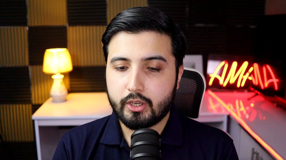 The image features a man wearing a blue shirt, sitting at a desk with a microphone in front of him. The background includes a neon sign that reads "Amana" along with some text about the current state of the gaming industry. The man appears to be speaking into the microphone or possibly recording something related to the gaming world.