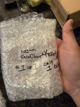 A hand holds a bubble-wrapped package with handwritten text reading "100mm Excellent Your Yatast" and "#1 of 1 Only 1 ex", likely a graded collectible card.