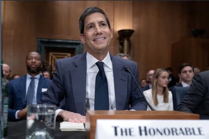 A man in a suit sits at a podium labeled "THE HONORABLE" with a microphone, smiling upward during what appears to be a congressional hearing, while attendees are seated behind him. The image likely relates to the Fed's planned Treasury bill purchases, suggesting the official pictured may be involved in announcing or discussing these market-injecting measures.