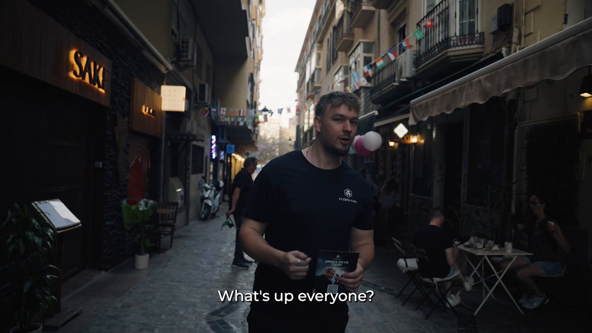 The image shows a man standing in an alleyway between two buildings with a sign that reads "Saqi". The man appears to be holding a pink balloon while looking at the camera. There are other people visible in the background, but they do not seem to be the main focus of the photo. The caption above the image suggests that this scene is part of an advertisement for the World Cup experience in Spain.