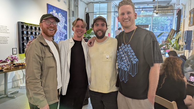 Four men pose together smiling for a photo indoors, with the man on the right wearing a black t-shirt featuring a blue graphic design.