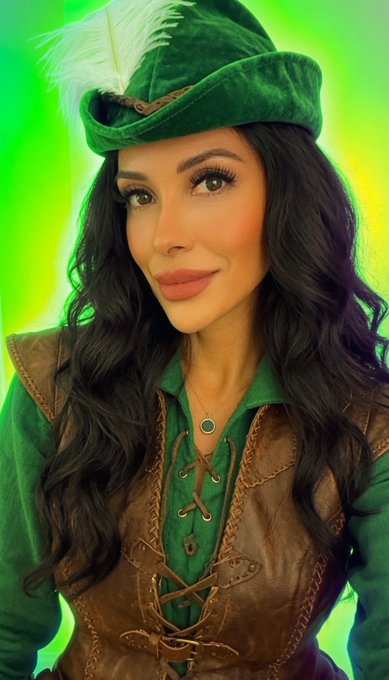 A woman with long dark hair wears a green velvet feathered hat and a brown leather corset with a green blouse, posing against a vibrant green and yellow background. The Robin Hood-inspired costume doesn't visually connect to the accompanying post about gem hunting and crypto research, which focuses on diligent analysis rather than the festive attire pictured.
