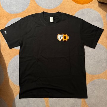 A black t-shirt featuring a "B" logo in gold and white on the chest is laid out on a patterned carpet, likely a promotional giveaway for the Cake Wallet booth at the Bitcoin Conference Asia event.