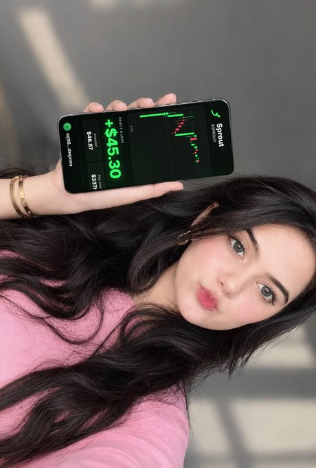 A young woman in a pink top holds up a smartphone displaying a green-trending trading chart on the Sprout app, with a value of +$45.30 visible on the screen.