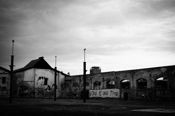 A black-and-white photo of a dilapidated industrial building covered in graffiti, featuring a prominent banner that reads 'NOW IS OUR TIME.'