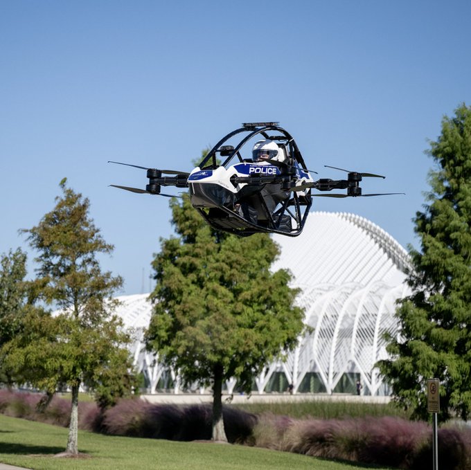A blue-and-white electric aircraft marked “POLICE” hovers over a park near a white domed structure, illustrating Florida Polytechnic’s debut of a manned flying police vehicle for surveillance and emergency response.