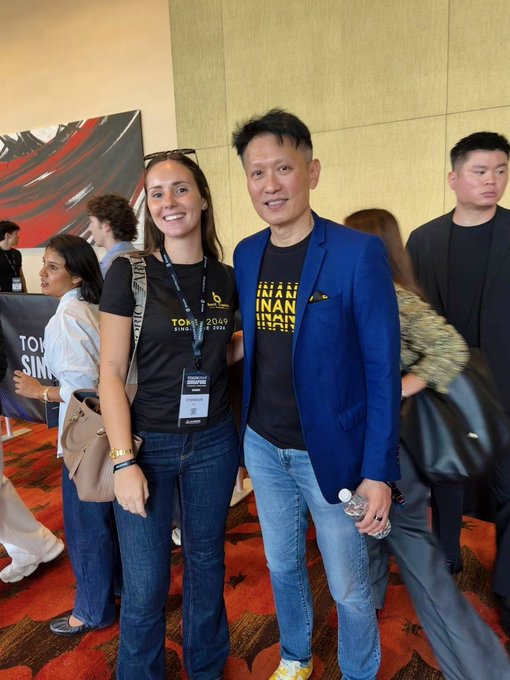 A woman wearing a "TOKEN 2049 SINGAPORE" lanyard and t-shirt poses for a photo with a man in a blue blazer featuring "FINANIN" branding, both smiling for the camera at the Token2049 Singapore event. The "TOKEN 2049" signage visible in the background confirms the conference setting referenced in the caption about running into someone on Day 2.