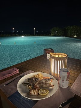 A nighttime photo shows a dining table by an infinity pool, featuring a plate of grilled seafood, a bread basket, an LED lantern, and a beverage can on a wooden surface.