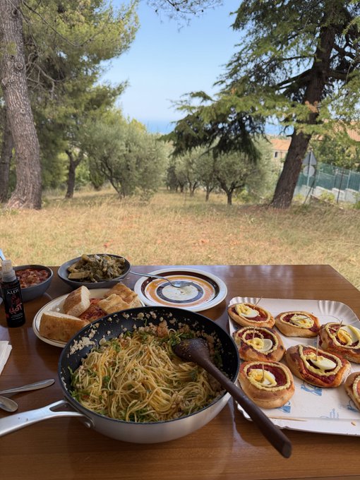 An array of homemade Italian-inspired dishes—spaghetti in a pan, bruschetta topped with eggs, bread, olives, and what appears to be jam—sits on a wooden table overlooking a sunlit olive grove with the sea in the distance. The post suggests a return to simple, traditional living after a digital distraction, with the rustic, outdoor meal symbolizing a focus on real, tangible pleasures.