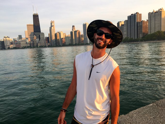 A smiling young man wearing sunglasses, a camo bucket hat, and a sleeveless white Nike shirt, posing in front of the Chicago skyline along Lake Michigan.