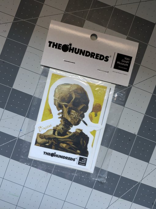 A packaged "THE HUNDREDS" x Van Gogh Museum sticker featuring Van Gogh's "Skull of a Skeleton with Burning Cigarette" artwork, resting on a grid cutting mat.