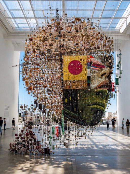 A large museum installation featuring a suspended face made of numerous small objects, displayed in a sunlit gallery with visitors viewing it.
