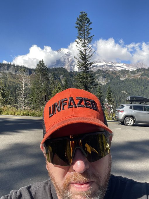 A man wearing an orange "UNFAZED" cap and reflective sunglasses takes a selfie in front of a majestic, snow-capped mountain (likely Mt. Rainier), with his parked car visible in the background. The cap's "UNFAZED" text visually echoes the post's message of staying calm and unbothered.