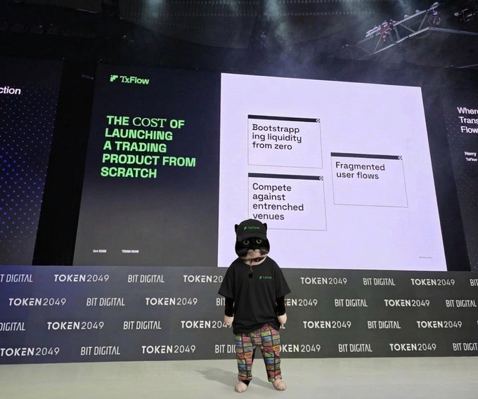 A person wearing a black cat-eared hat and colorful patchwork pants stands barefoot on a stage at TOKEN2049 BIT DIGITAL, in front of a presentation screen titled 'THE COST OF LAUNCHING A TRADING PRODUCT FROM SCRATCH' by TxFlow.