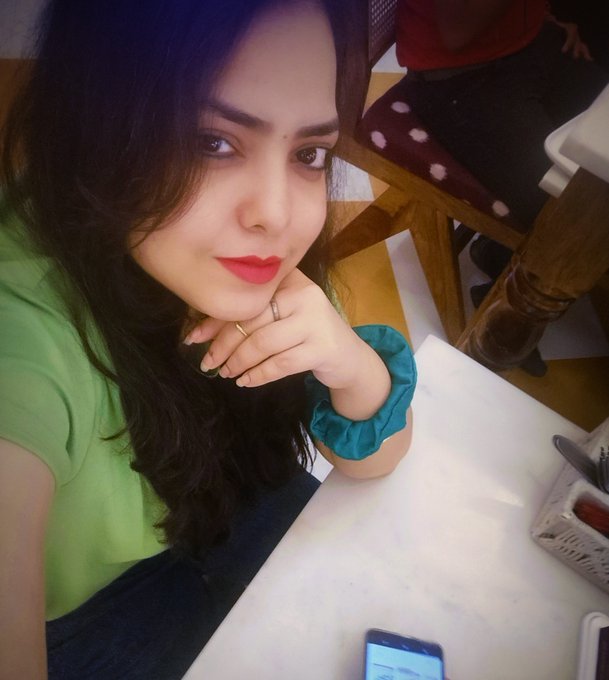 A selfie of a woman with long dark wavy hair, wearing a green top and red lipstick, resting her chin on her hand which displays two rings; a smartphone is visible on the white table beside her.