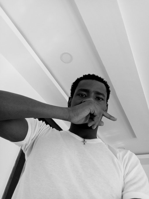 A black-and-white low-angle selfie of a young man wearing a white t-shirt and a cross necklace, holding his hand across his face.