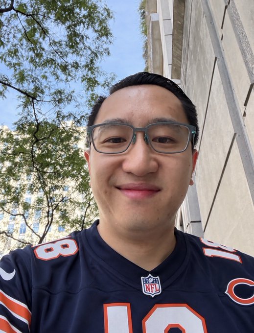A selfie of a smiling man wearing glasses and a navy blue Chicago Bears NFL jersey (number 18) taken outdoors with trees and a stone building in the background.