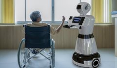An elderly person in a wheelchair interacting with a white humanoid robot in a bright indoor space.