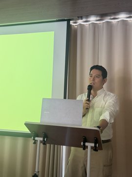 A man in a white shirt stands at a podium holding a microphone, presenting in front of a green projection screen during the Hermes agent session where Andrea Klaydice explained x402 use cases.