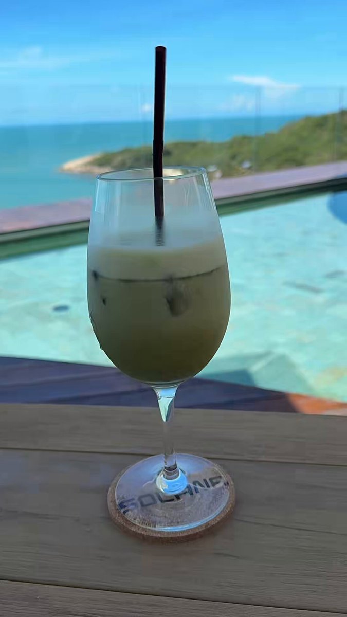 The image features a glass of green tea sitting on top of a coaster, with a straw sticking out of it. The cup has a label that reads "Solana summer", suggesting that this drink might be inspired by the summer season or associated with a specific location. The setting appears to be outdoors, possibly at a poolside or beachfront area, as there is a view of water in the background.