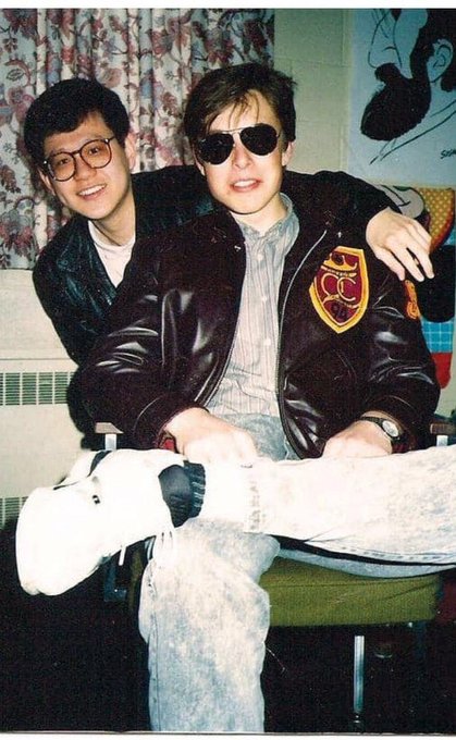 A vintage photograph shows two young men posing closely together indoors, one wearing a leather jacket with a yellow shield patch and dark sunglasses while seated, and the other standing behind him with glasses and a friendly smile. The retro styling and casual pose match the post's nostalgic caption referencing Elon (likely Elon Musk) and "CZ" (Changpeng Zhao) before they became well-known tech figures, suggesting a youthful "old school" moment between the two before they went on to transform their respective industries.