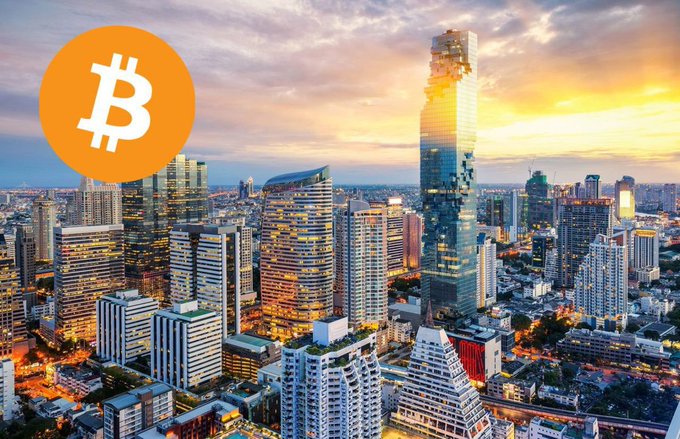 A city skyline at sunset with a Bitcoin (₿) logo overlaid in the upper left corner.