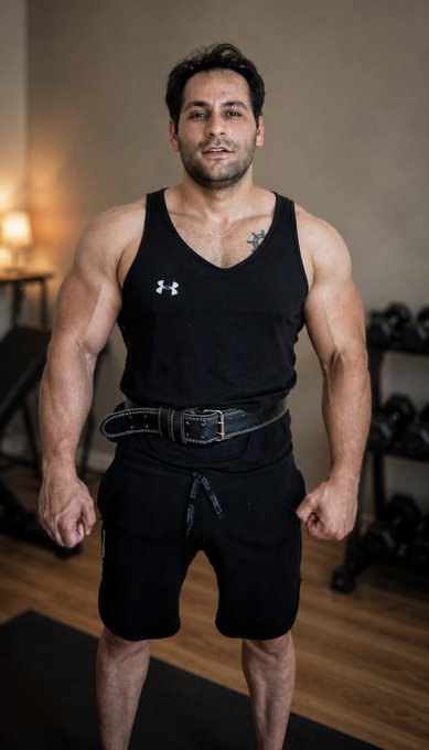 A muscular man wearing a black Under Armour tank top and black shorts with a weightlifting belt, standing in a gym with dumbbells in the background.