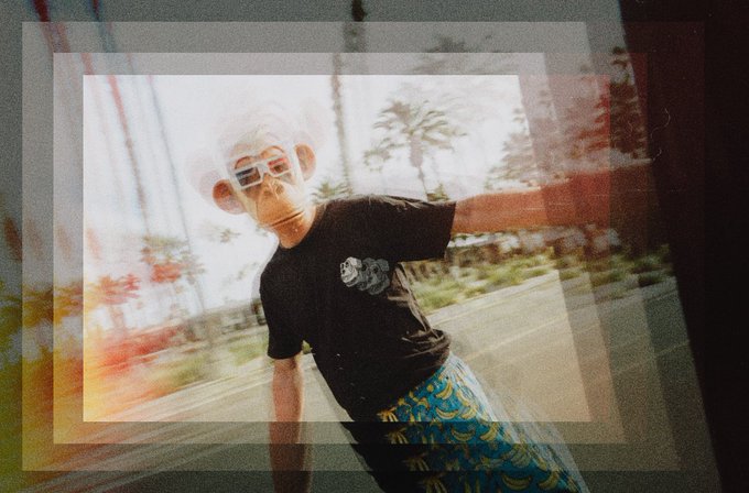 The image features a person wearing sunglasses, a black t-shirt with a monkey mask, and blue shorts. They are standing outdoors, possibly at an amusement park or a themed event. The photo has been edited using a filter that gives it a bokeh effect, making the subject appear as if they have a face. The caption of the image reads "GM focus! bored ape yacht club". This suggests that the person in the picture is wearing a funny and eye-catching costume to draw attention or participate in an event related to the "GM Focus!" theme.