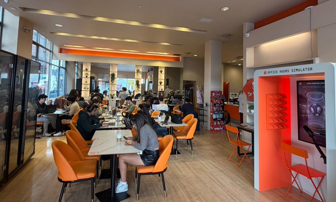 Customers dine at orange tables inside a brightly lit, bustling cafe while an "Office Hours Simulator" interactive booth stands prominently to the right, illustrating the company's open and operational locations amid the temporary closure controversy.