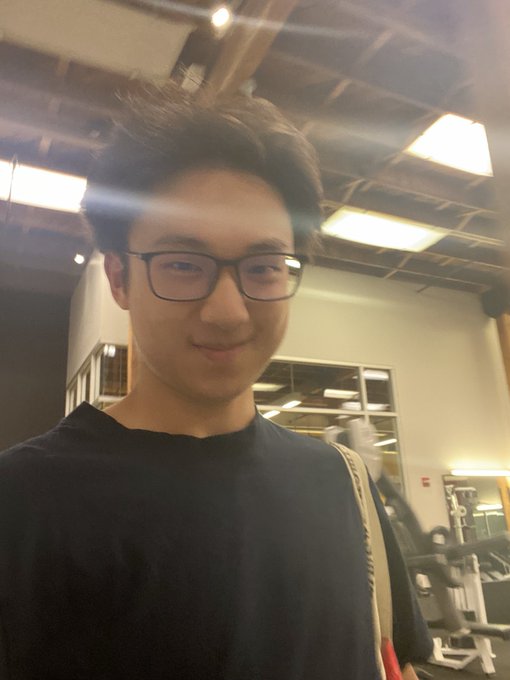 A young man with glasses and a black t-shirt poses for a selfie in what appears to be a gym, with weight equipment visible behind him, while his caption humorously suggests he could fit the @o1_exchange platform into the space.
