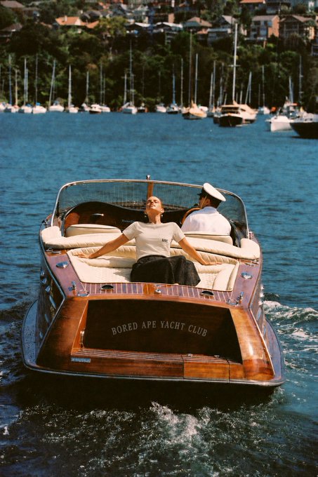 A woman in a white t-shirt lounges with arms outstretched on a wooden speedboat as a man in a captain's hat steers through a harbor of moored sailboats. The boat's stern prominently displays the text "BORED APE YACHT CLUB" — a literal and playful nod to the BAYC reference, blending luxury yacht culture with the iconic NFT brand.