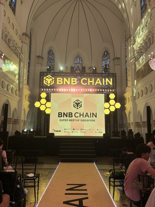 An ornate cathedral-like space hosts the "BNB CHAIN SUPER MEETUP SINGAPORE" event, featuring a stage with the BNB Chain logo and a lit runway leading to the stage.
