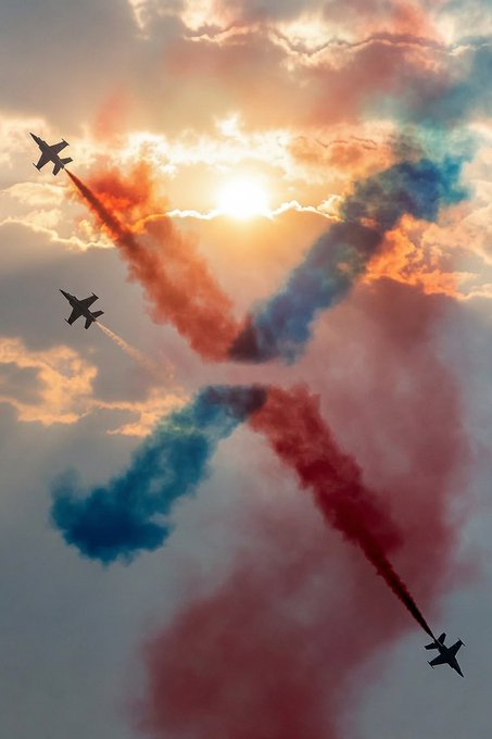 Aerial photograph of jets performing an airshow with red and blue smoke trails crossing in an X pattern against a sunlit cloudy sky.