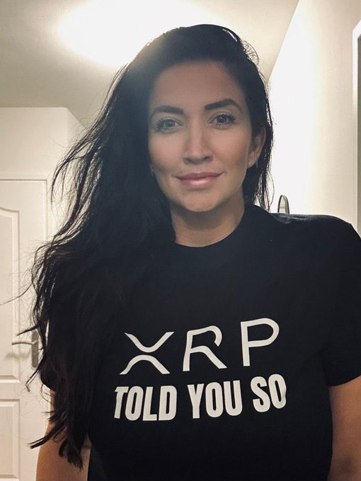 The image features a woman with long hair wearing a black t-shirt that has white text on it, which reads "told you so" followed by the phrase "XRP". The background of the photo shows a bathroom setting with a sink visible in the scene.