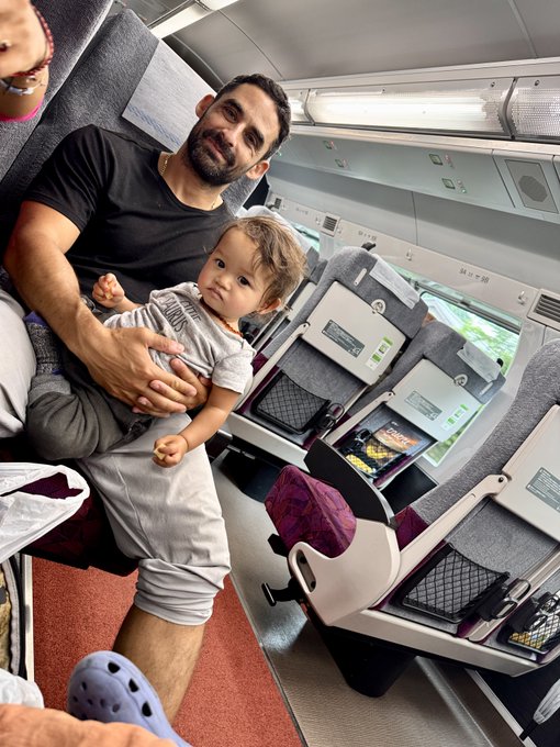 A bearded man sits on a train holding a young child on his lap, with luggage and a Crocs shoe visible nearby. The image accompanies a personal account about discovering $1.6M in Bitcoin was stolen, contrasting a calm, composed demeanor with the shock of a million-dollar theft.