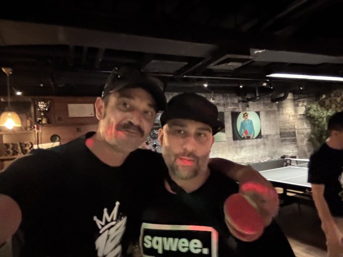 Two men wearing black caps pose together in what appears to be a dimly lit bar or restaurant, with a ping pong table visible in the background.