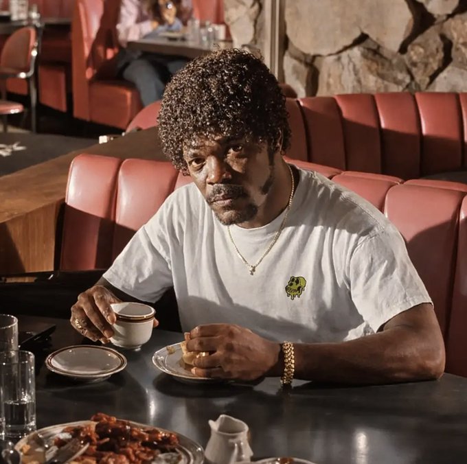 A man with a curly afro sits in a diner booth wearing a white t-shirt, gold chain, and gold bracelet, holding a coffee cup with breakfast on the table. The casual diner setting and the man's direct, deadpan gaze into the camera match the irreverent, tongue-in-cheek energy of "Happy Mutant Monday Motherfucka."