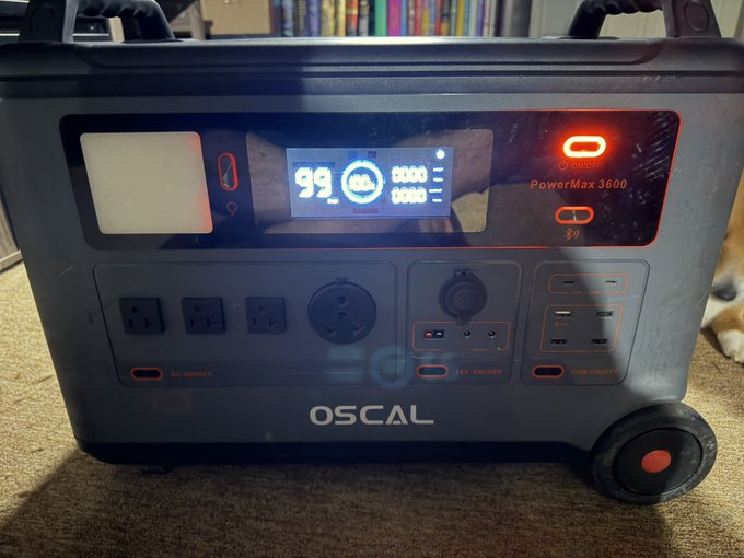 An OSCAL PowerMax 3600 portable power station displays a 99% charge on its digital screen, featuring various outlets, switches, and an illuminated power button.