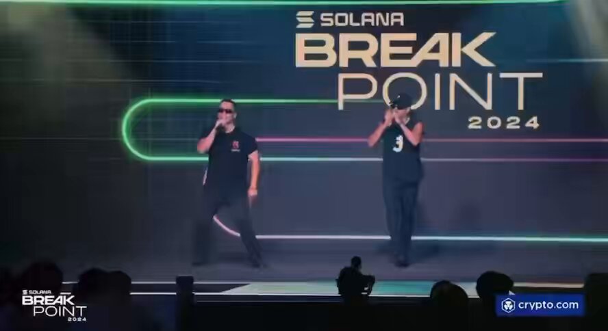 The image features two men performing a dance routine on stage, with one man wearing sunglasses and holding a microphone while the other has his hands up in the air. They are standing next to each other, showcasing their talent for dancing. The background of the image is black, which contrasts with the colorful neon lights that illuminate the stage. The text "solana summer" can be seen on the image, possibly indicating an event or promotion related to the Solana cryptocurrency platform.