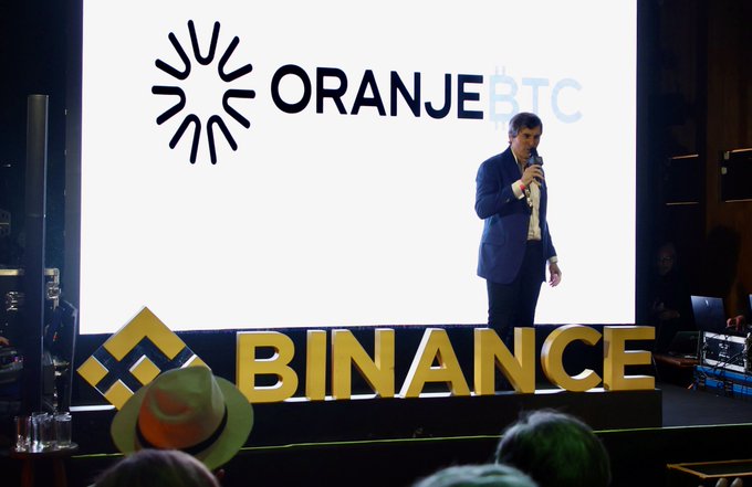 A man in a suit speaks into a microphone on stage at a 'BINANCE' event, with a large screen behind him displaying the 'ORANJE BTC' logo.