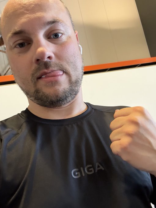 A close-up selfie of a bearded man wearing wireless earbuds and a black "GIGA" t-shirt, making a fist gesture in what appears to be a gym or fitness facility.