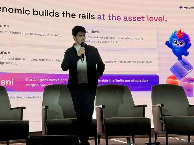 A young man in a black jacket speaks into a microphone on a stage with empty chairs in the foreground, standing in front of a presentation screen with text including 'nomics builds the rails at the asset level' and 'Validate', alongside a blue and pink cartoon mascot character from 'Kenomist'.