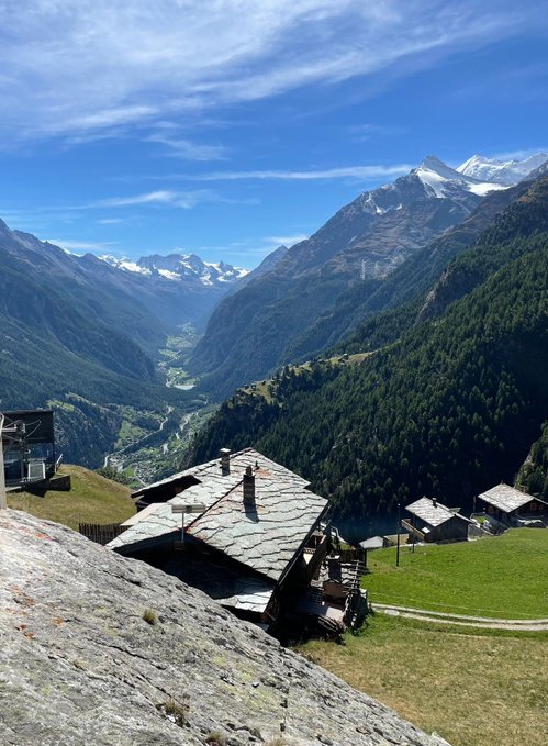 A scenic mountain landscape featuring snow-capped peaks in the distance, a deep green forested valley, and traditional alpine stone chalets in the foreground under a clear blue sky.